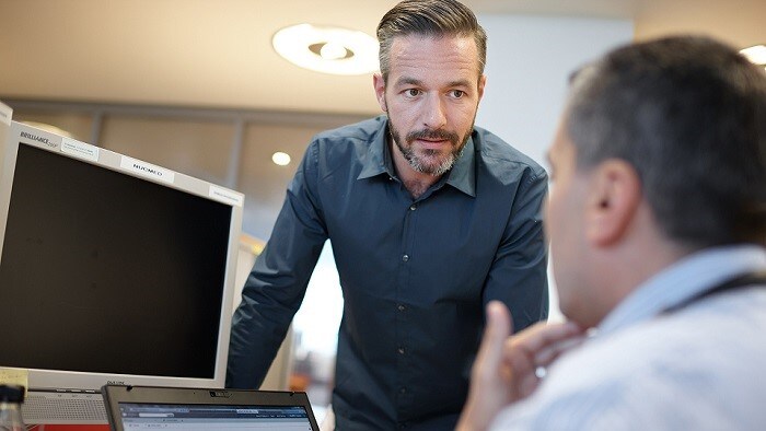 Two men have a conversation at a desk in front of a monitor