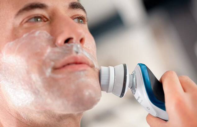 Nærbillede af en mand med barberskum i ansigtet. Han bruger en hvid, blå og grå elektrisk barbermaskine.
