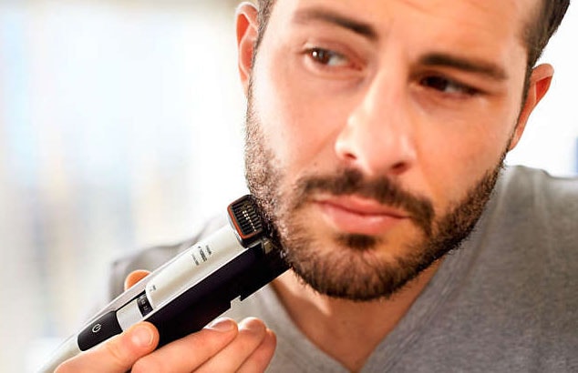 Nærbillede af en mørkhåret mand med skæg i en grå T-shirt, som bruger en elektrisk barbermaskine på sin kæbelinje.
