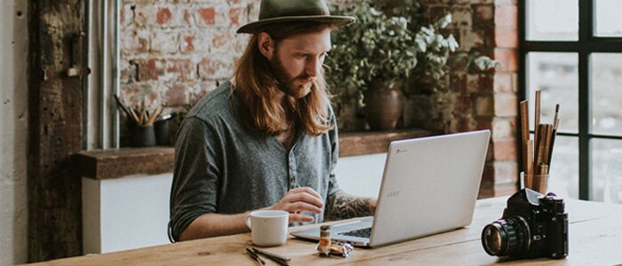 En ung mand med langt hår og fuldskæg sidder et hipt sted med en laptop foran sig.