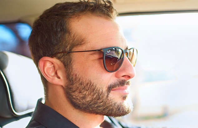 En mand i en bil og med sorte solbriller på har et brunt fuldskæg, overskæg og kortklippet hår.