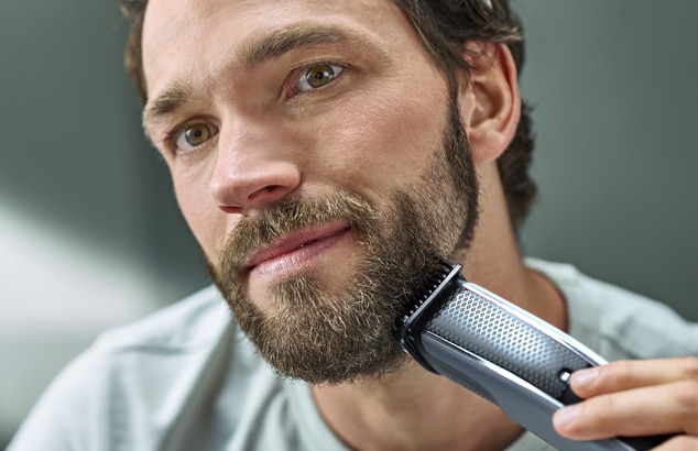 En mand med et fuldskæg barberer sit ansigt med en sølv- og sortfarvet elektrisk barbermaskine.