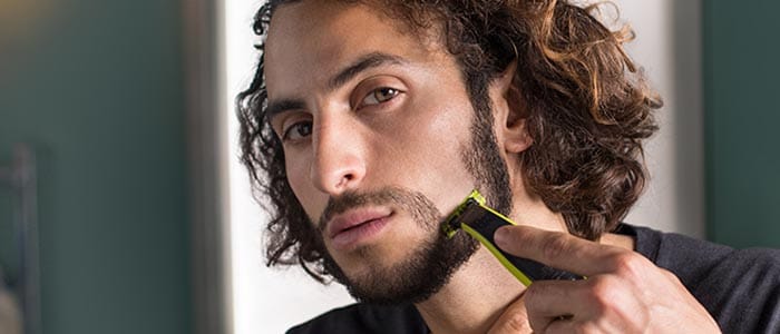 En langhåret mand bruger en trimmer til at barbere hele sit skæg og forme sit overskæg.