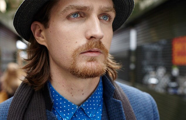 Mand med skulderlangt hår og Henriquatre-skæg bærer en blå skjorte, en blå blazer og en hat.