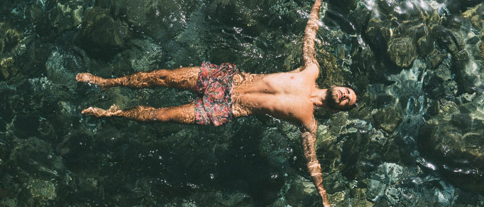 Et bredt billede af en skægget mand i svømmeshorts, som flyder i en pool på ryggen med sine arme strakt ud til siden.