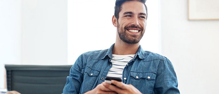 En mand iført skjorte med stubbet skæg holder en mobiltelefon mens han smiler.