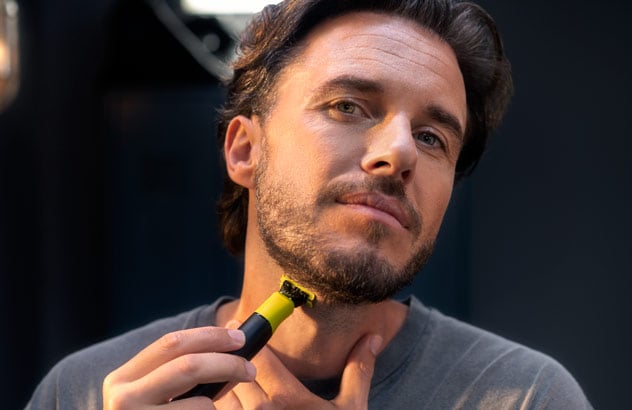 Nærbillede af en mand, som trimmer kanten af sit mørke skæg med en Philips OneBlade foran et spejl