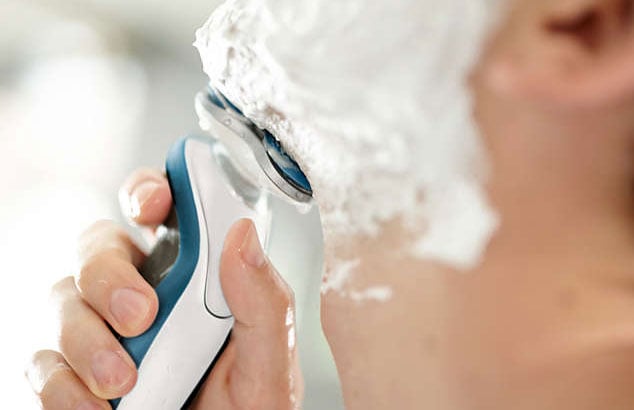 Nærbillede af en mand, som barberer sig med en hvid og blå elektrisk barbermaskine, hvor hagen er sløret og barbermaskinen er i fokus.