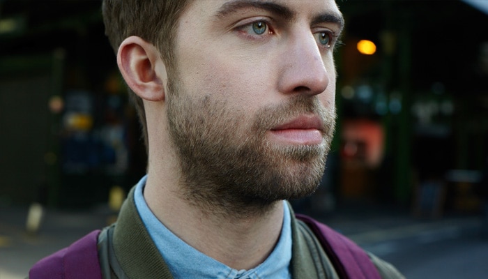 Brunhåret mand med 1 cm langt skæg, som dækker ansigt og nakke, står på gaden iført en lilla rygsæk. 
