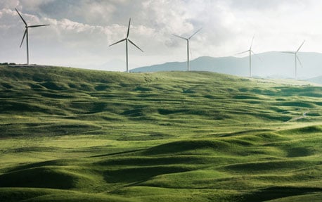 Et billede af en grøn skråning med roterende vindmøller, der genererer vedvarende energi, i baggrunden.